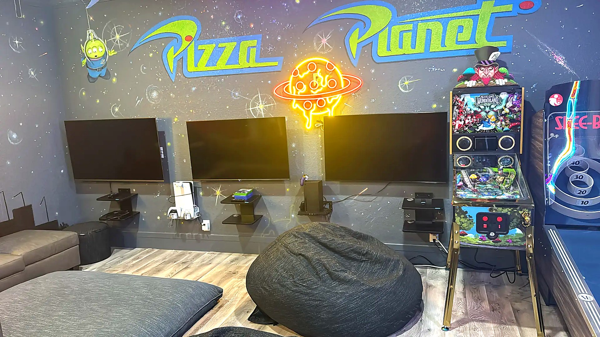 Game room gaming wall with three TVs, consoles, and Alice in Wonderland pinball machine